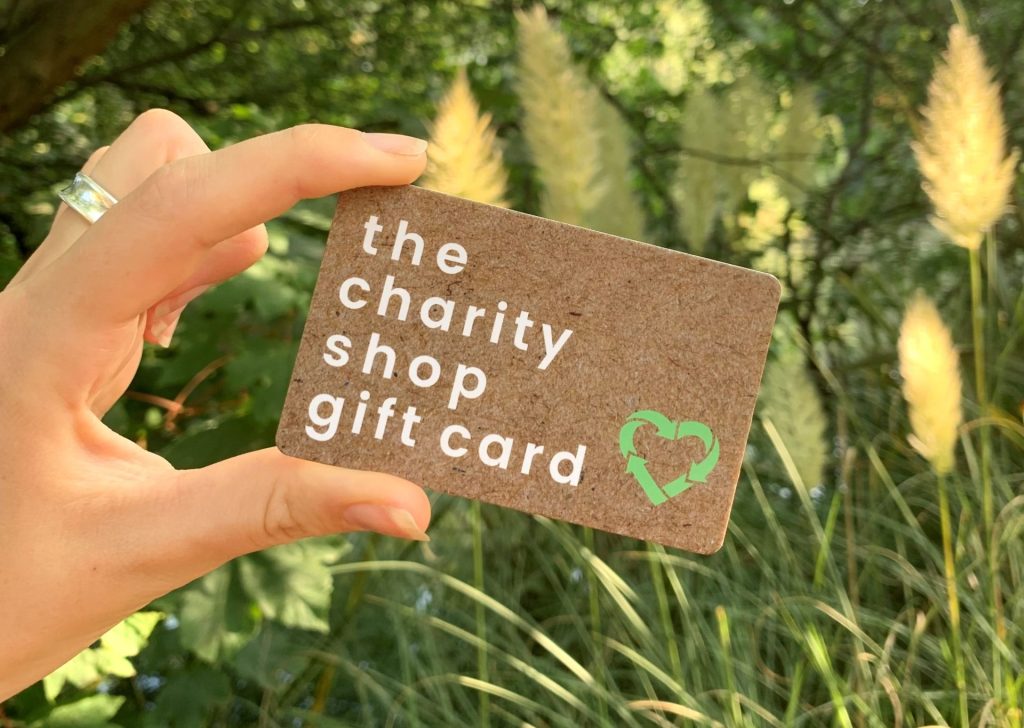 Charity Gift Card scheme ladies hand holding a recyclable brown charity shop gift card against a backdrop of trees and grasses