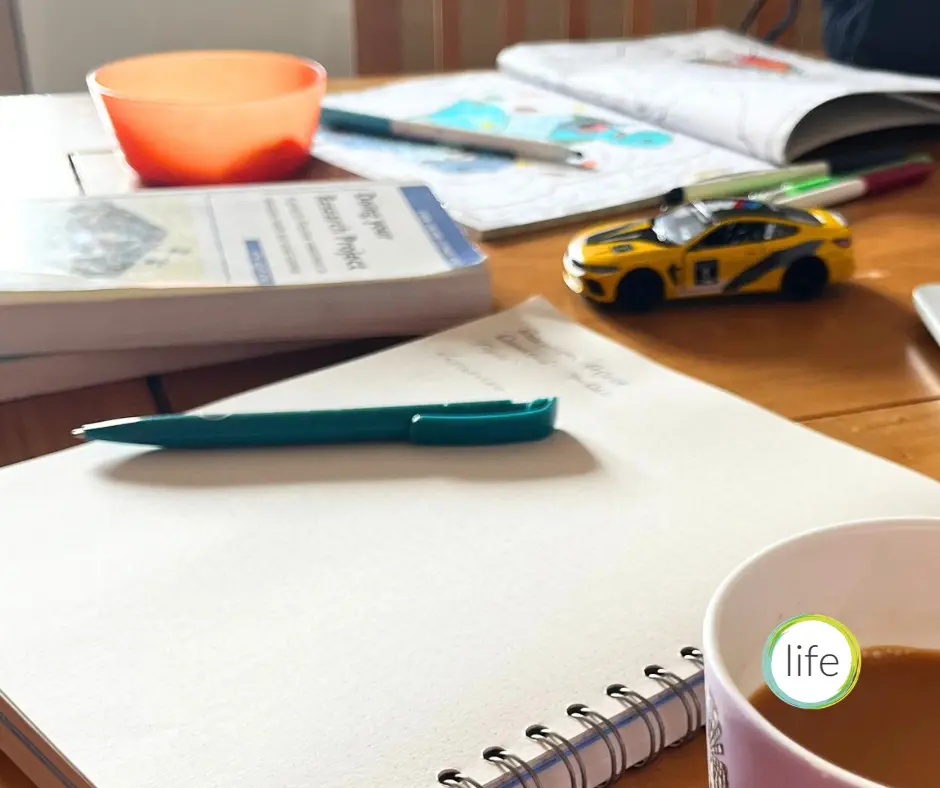 Busy kitchen table with a cup of tea, study materials and children's toys and cups